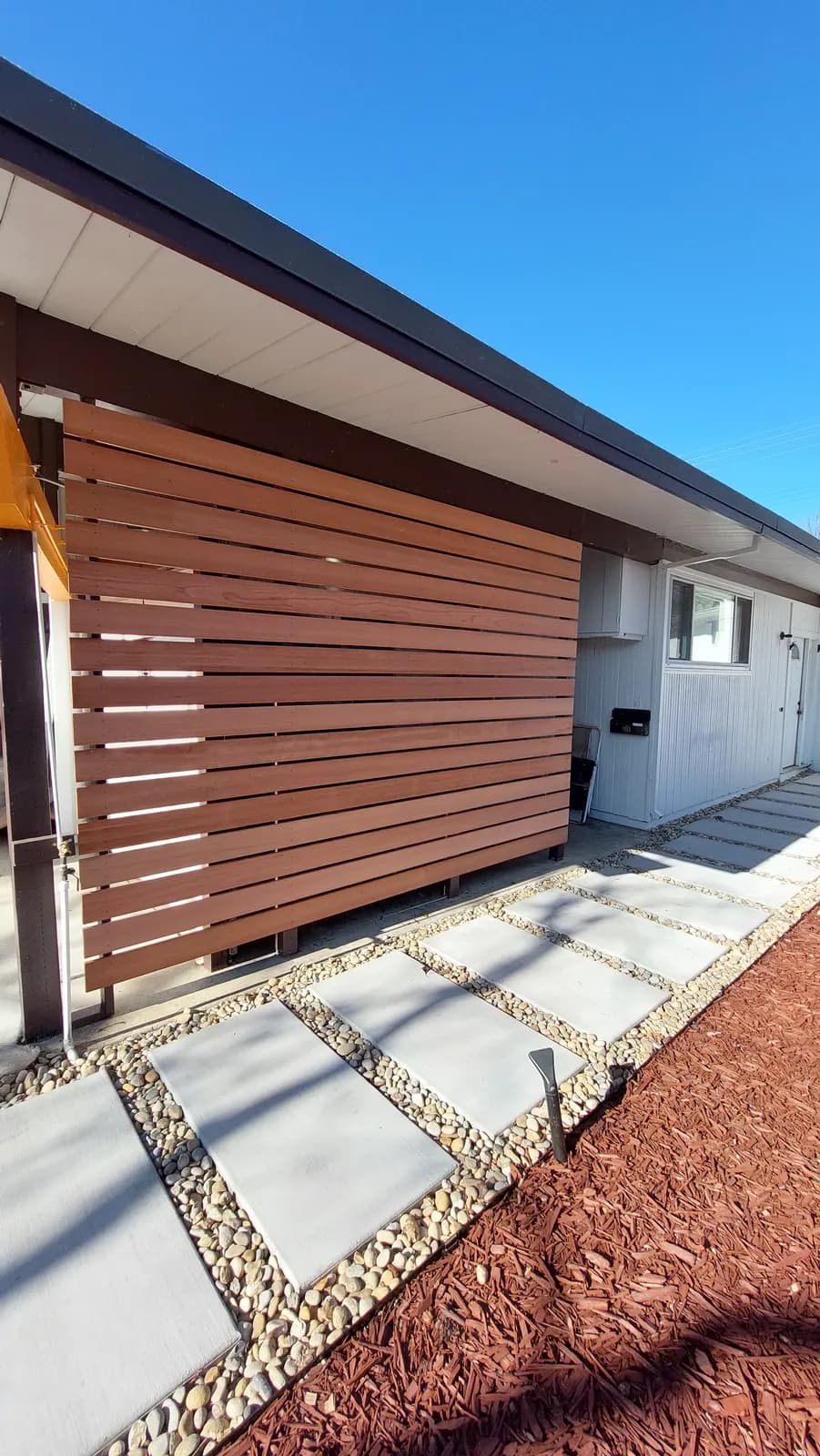 Horizontal redwood slat screen between posts under a flat roof overhang, paver and pebble path running past it
