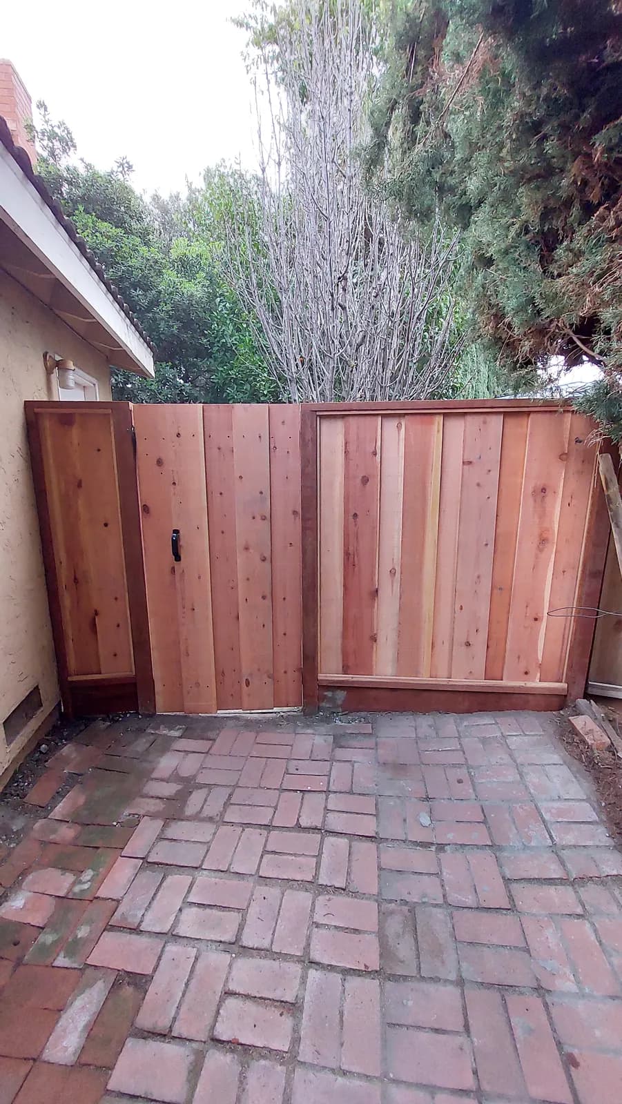 Redwood gate with a black pull handle beside a matching fence section, old brick paving underfoot