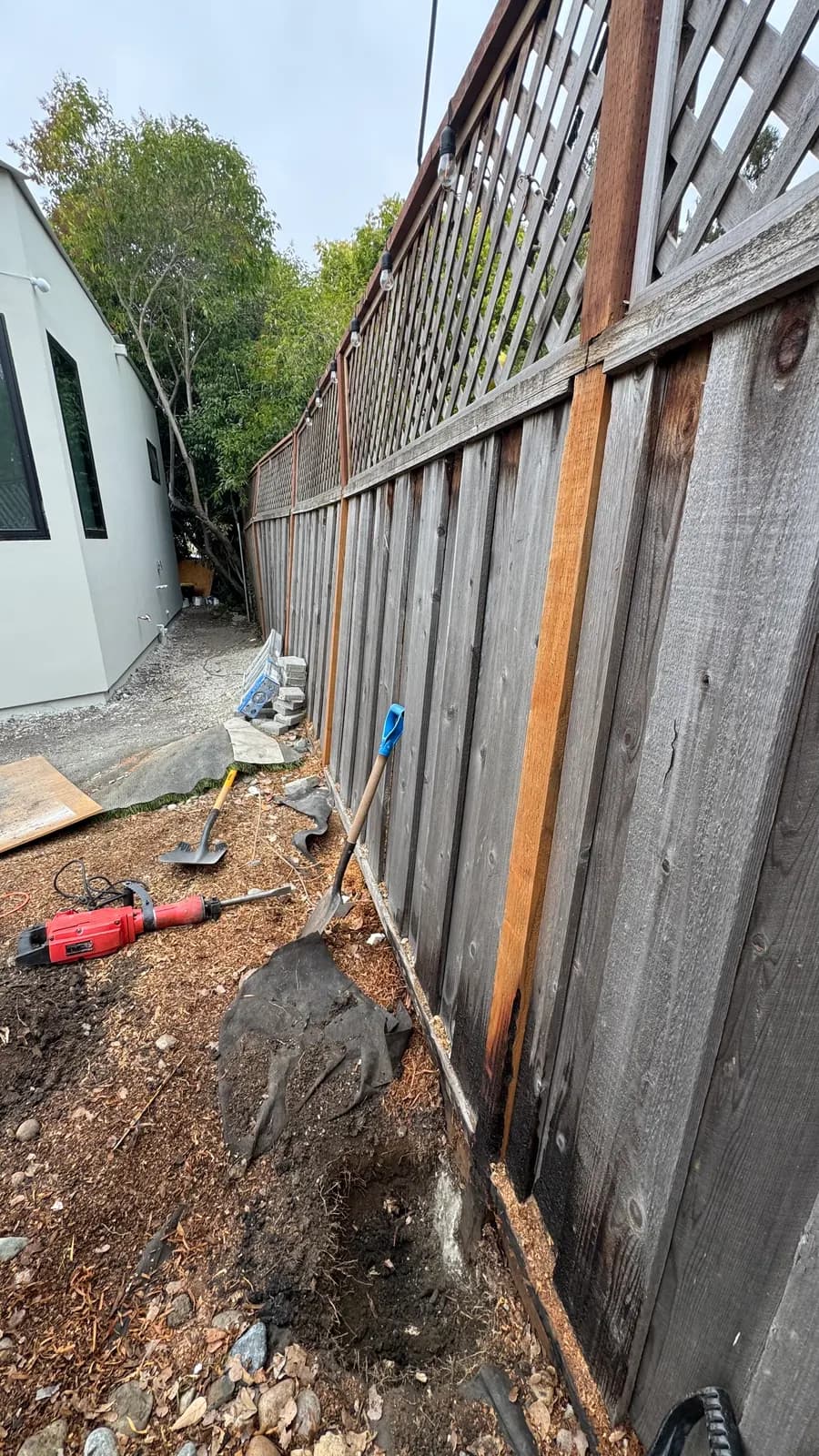 Long weathered fence with lattice top, one new red post already installed, shovels and a breaker on the opened ground