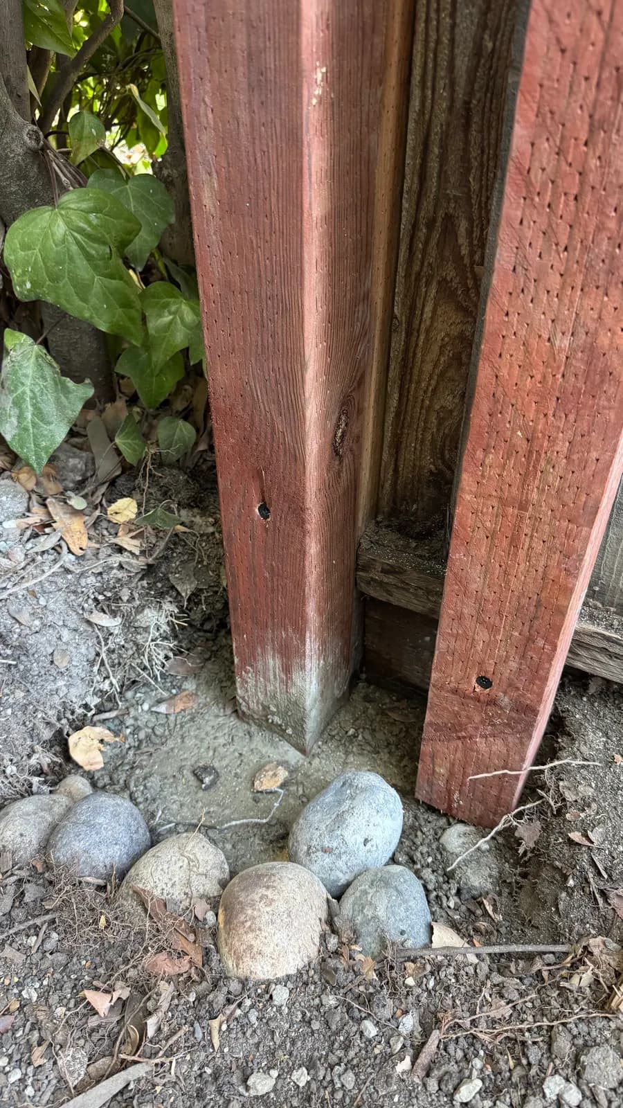 New redwood fence post set in a crowned concrete collar with river stones around it, weathered grey boards behind